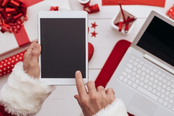 santa holding tablet on christmas surrounded by gifts